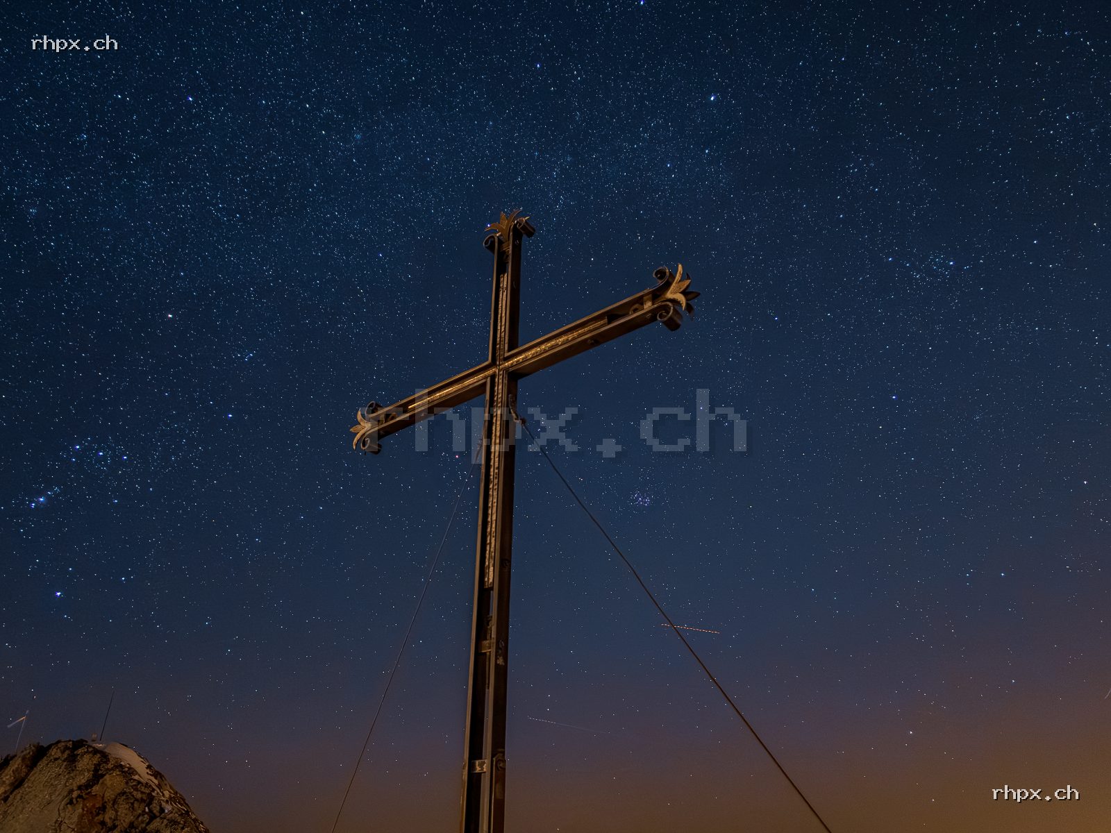 Illuminated summit cross on Mount Pilatus at night against a clear starry sky with an orange horizon