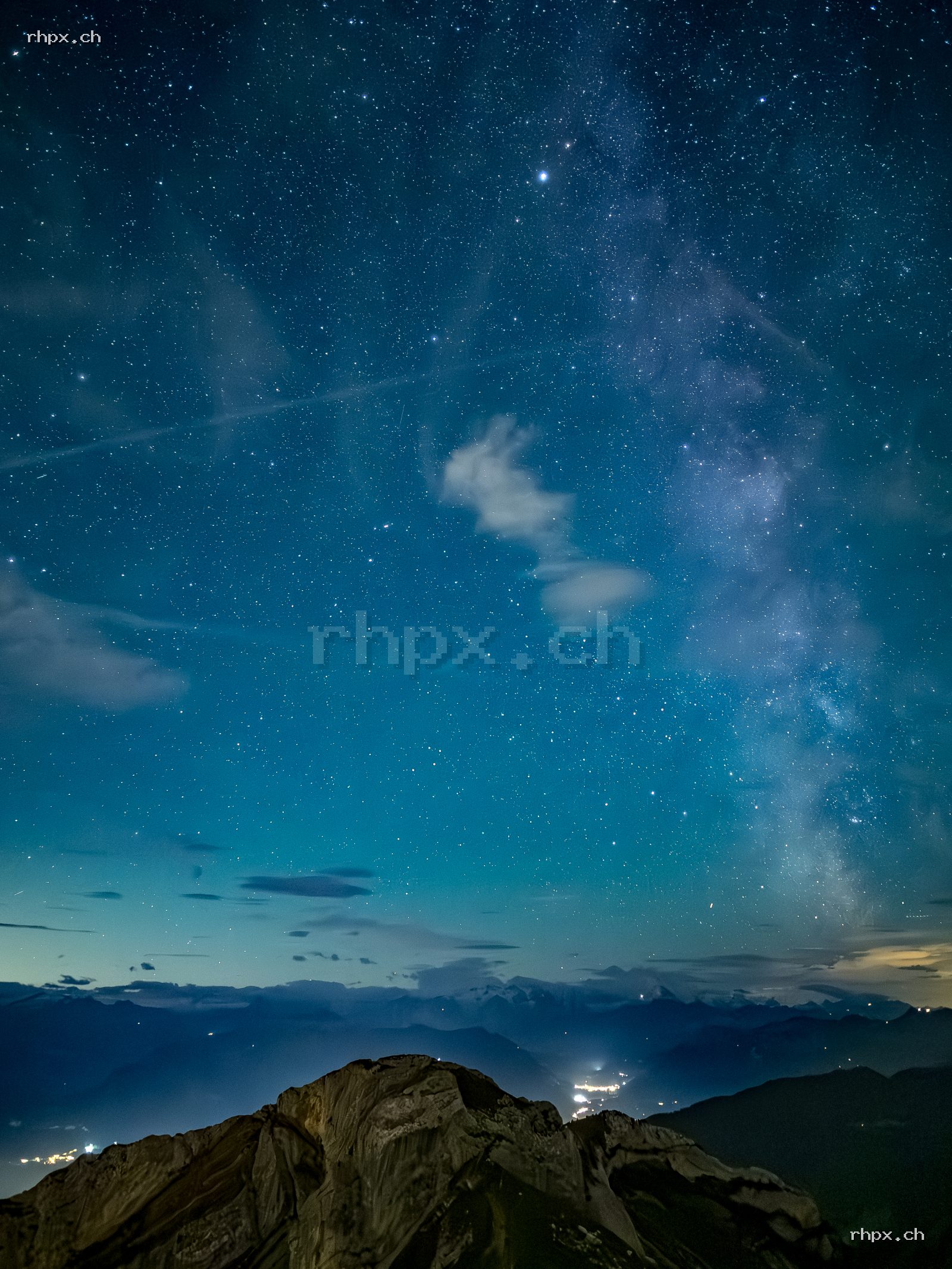 Milky Way and starry sky above a mountain panorama at night, rocks in the foreground, valley lights and clouds visible