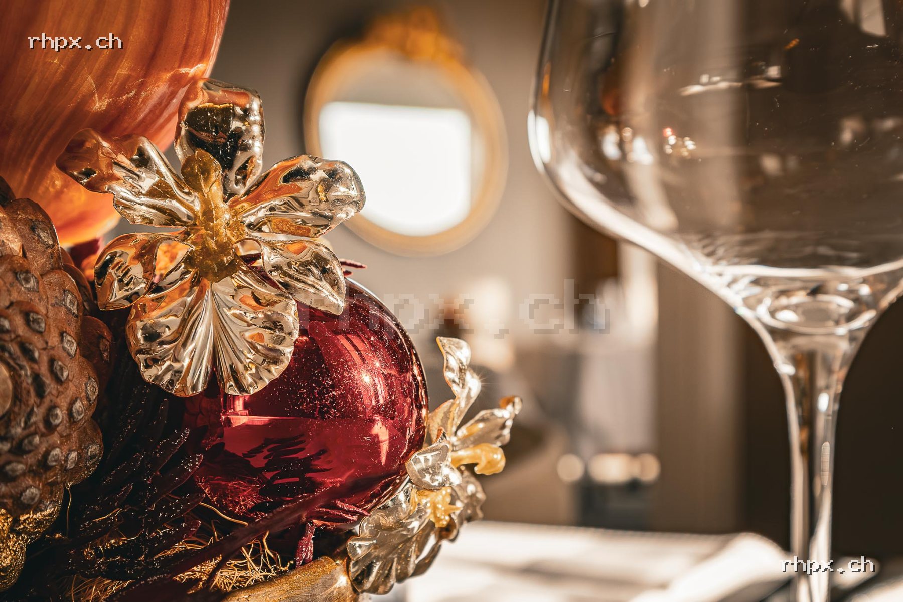 Close-up of festive decoration with a red ornament, golden flower, pine cone and wine glass in the background.