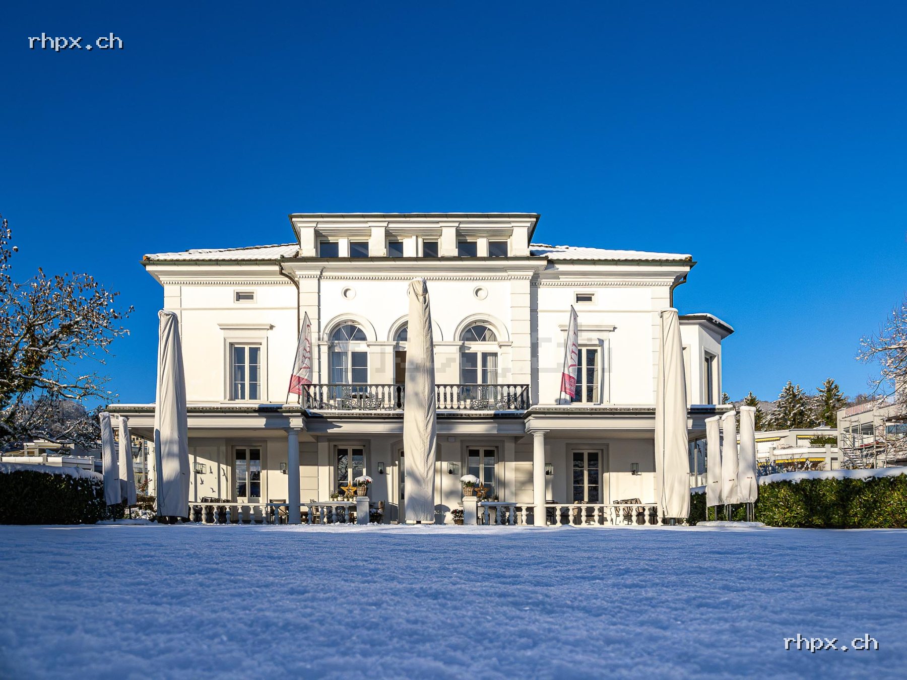 Front view of Villa Schweizerhof in winter with white facade, snow-covered lawn, closed parasols and blue sky.