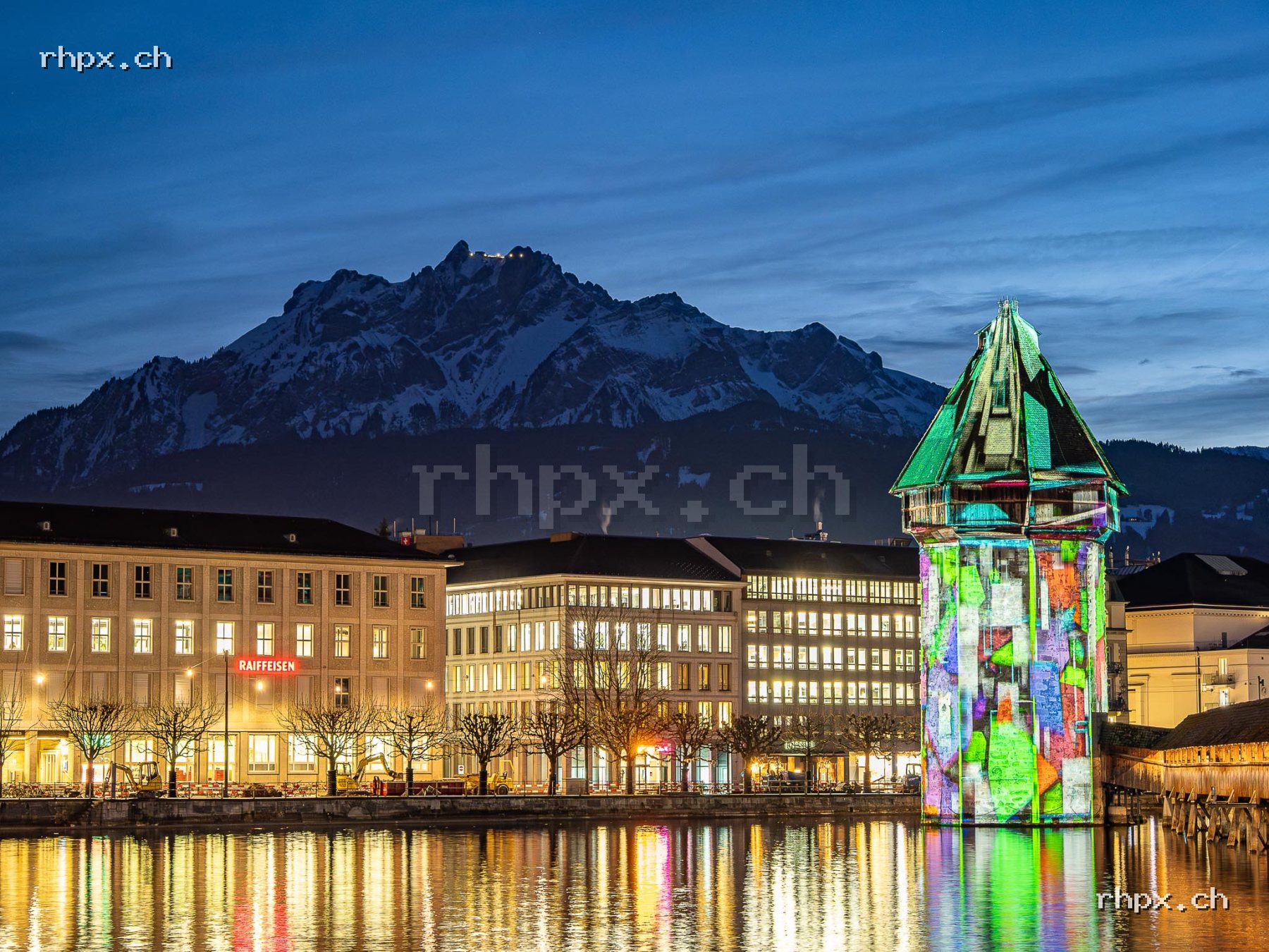Water Tower in Lucerne with colourful light projection, reflection in the Reuss and Mount Pilatus in the background.