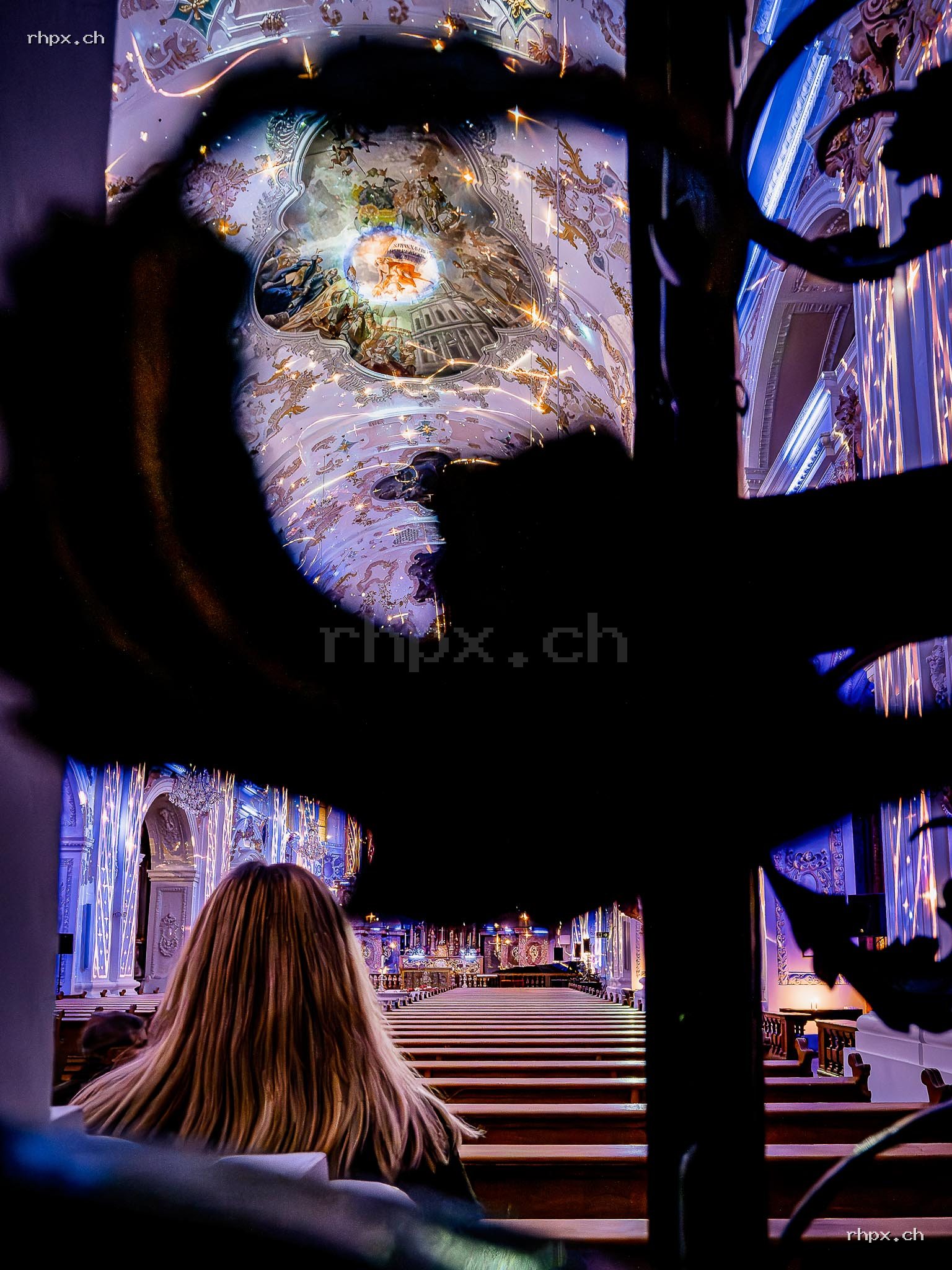 Person sitting inside the Jesuit Church looking towards a colourfully illuminated light installation in the church interior.