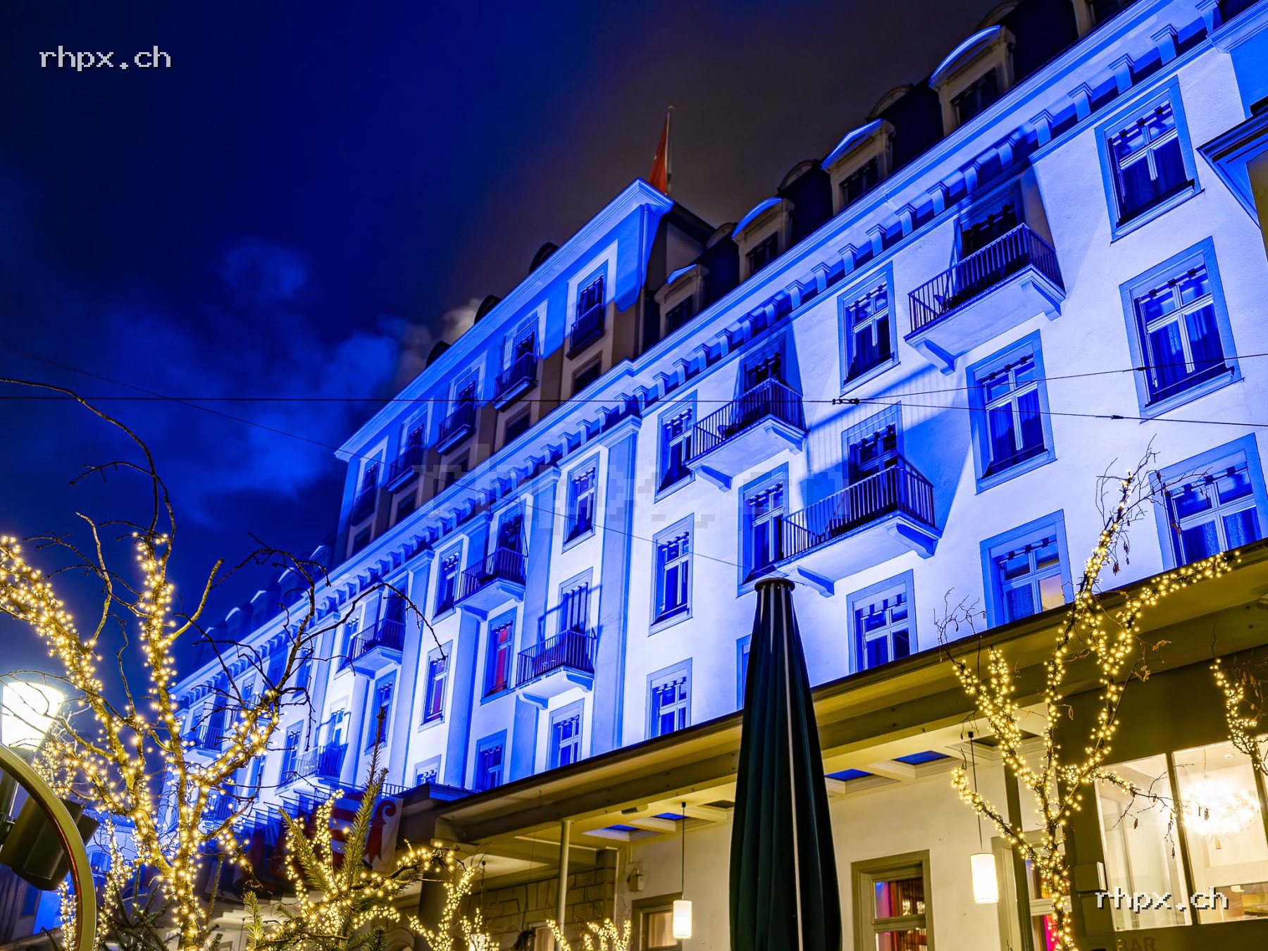 Facade of Hotel Schweizerhof Lucerne at night with blue lighting and illuminated trees in the foreground.