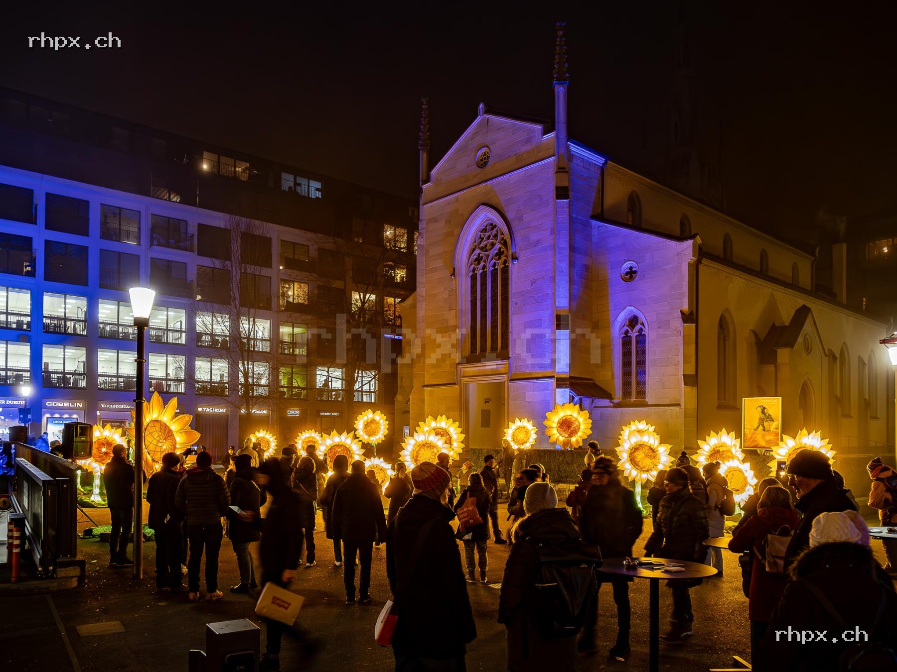 Illuminated flower installation in front of a church in Lucerne with many people in public space at night.