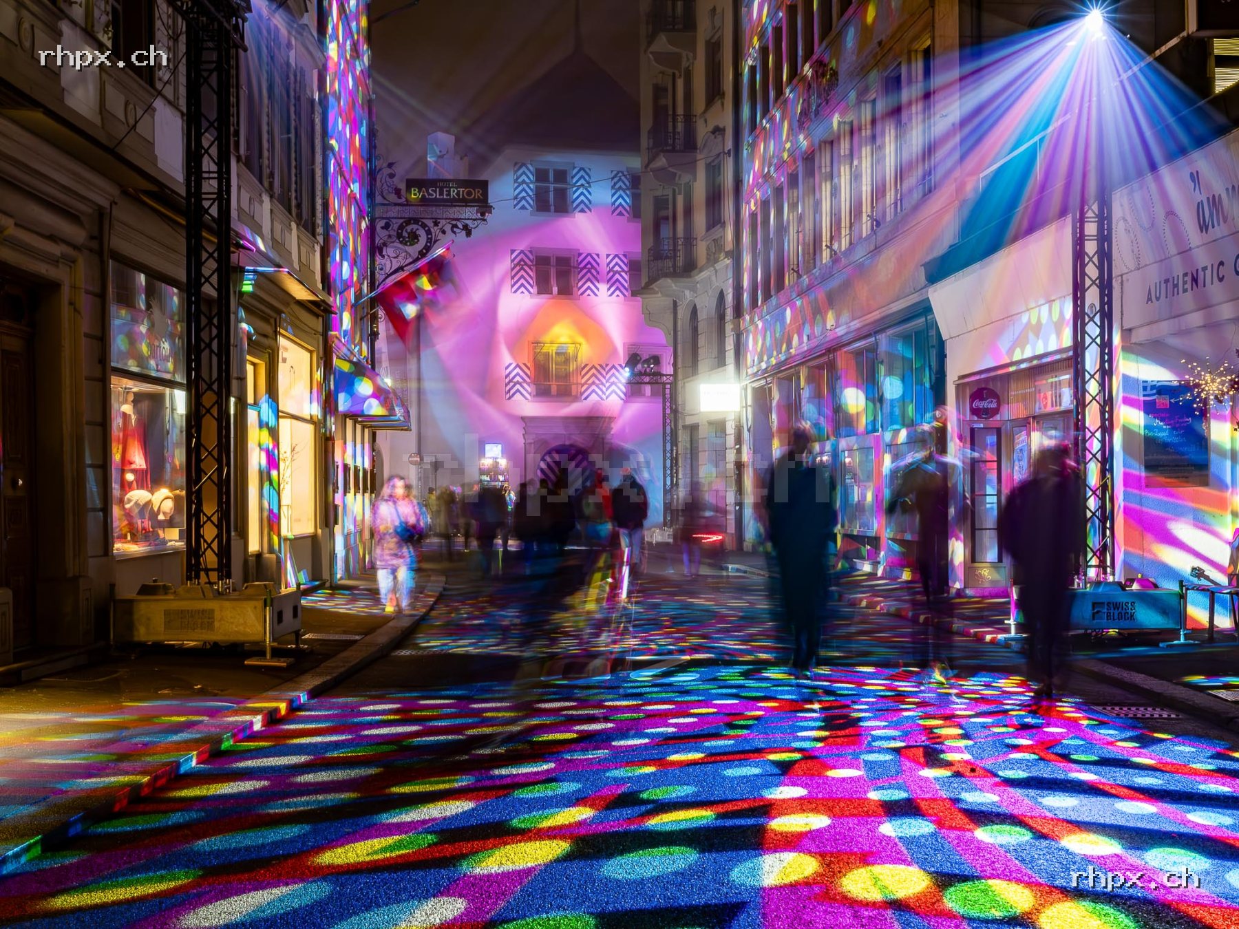 Old town street in Lucerne at night with colourful light projections on the ground and facades and people in the urban space.