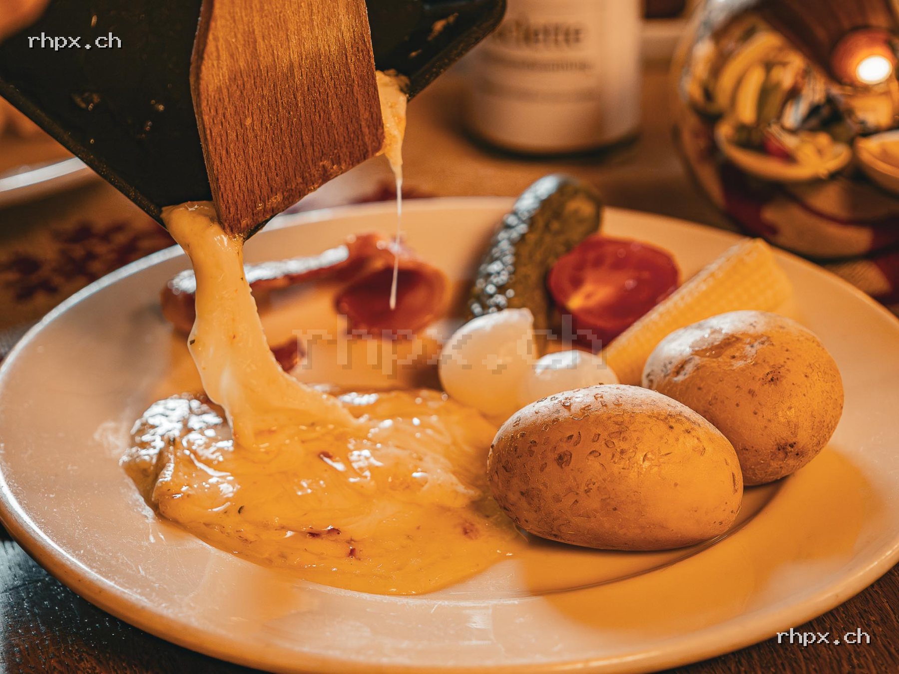 Melted raclette cheese is poured onto a plate with potatoes, pickles, pearl onions, bacon and corn.