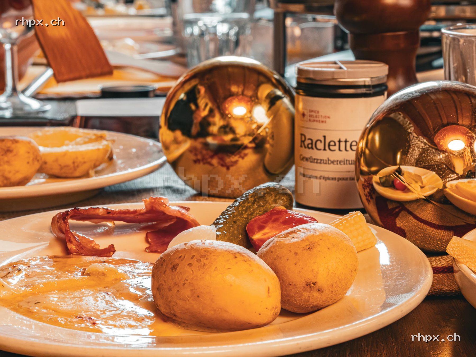 Raclette plate with potatoes, pickles, bacon and melted cheese on a laid table with additional ingredients and decoration.