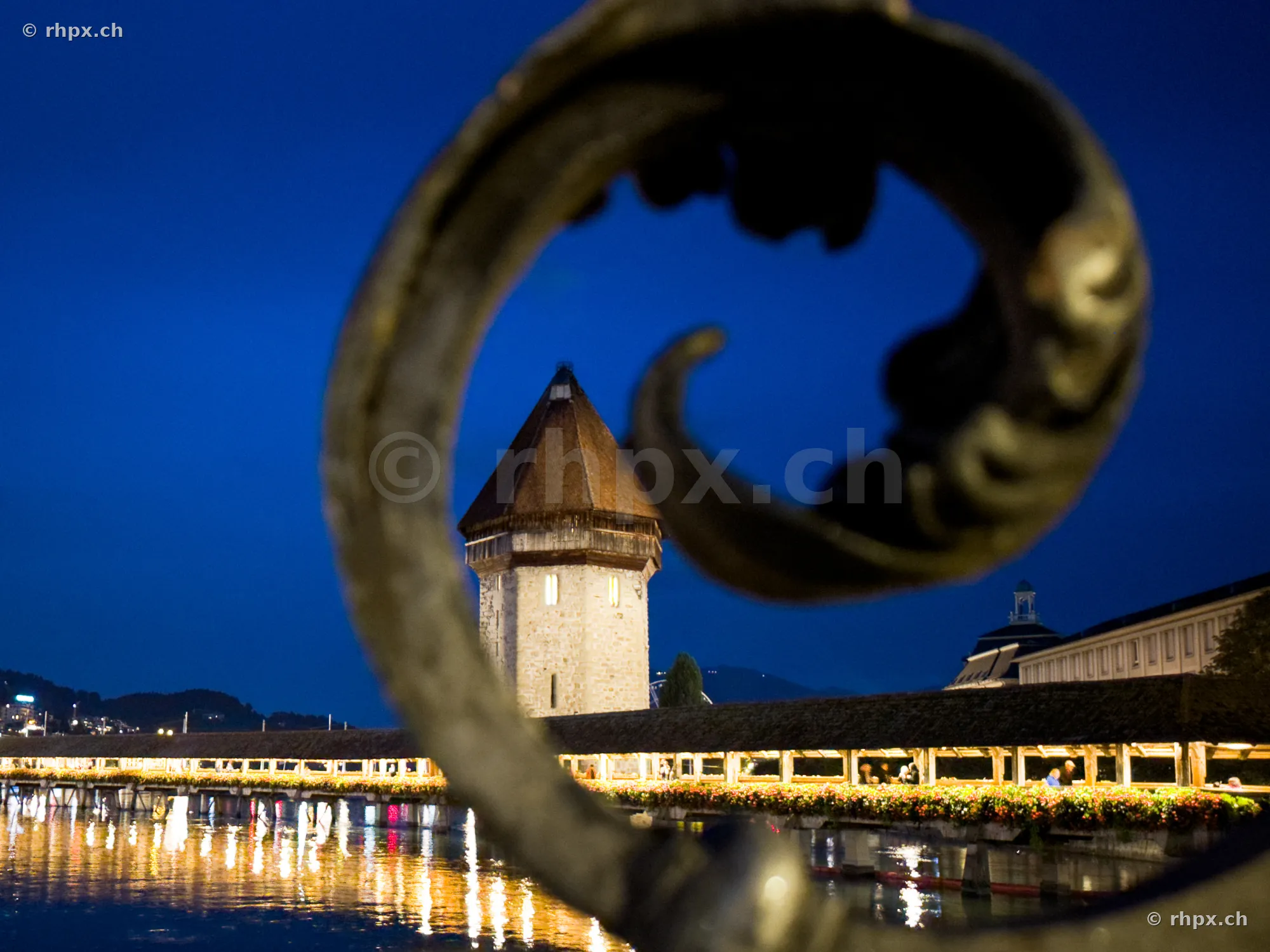 The Wasserturm and part of the illuminated Chapel Bridge in Lucerne at night, viewed through an ornamental iron railing.