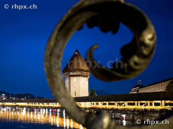 Der Wasserturm und ein Teil der beleuchteten Kapellbrücke in Luzern bei Nacht, betrachtet durch ein ornamentales Eisengeländer.