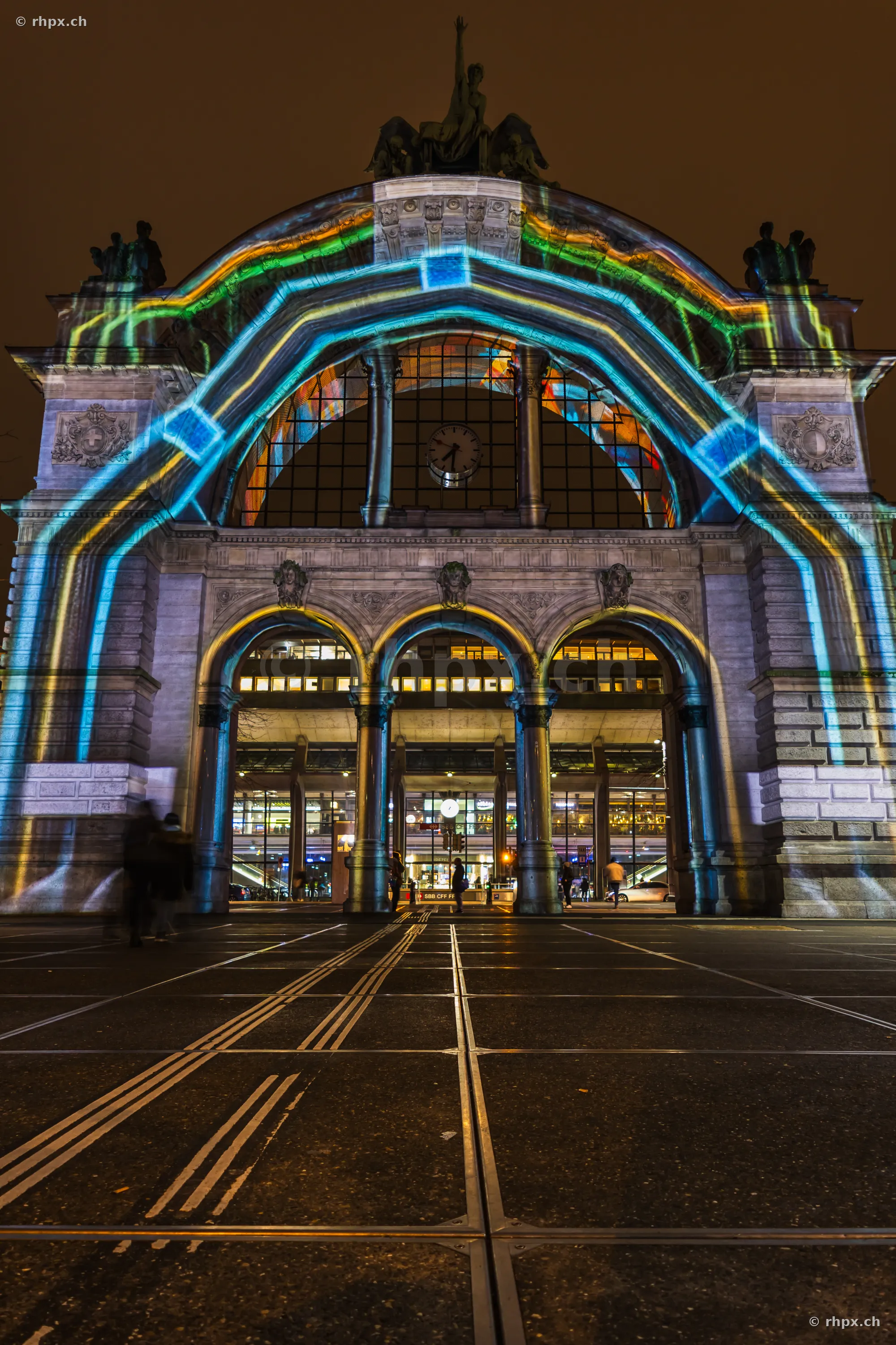 Fassade des Bahnhofs Luzern mit einer farbigen Lichtprojektion des Lilu Lichtfestivals 2024 bei Nacht.
