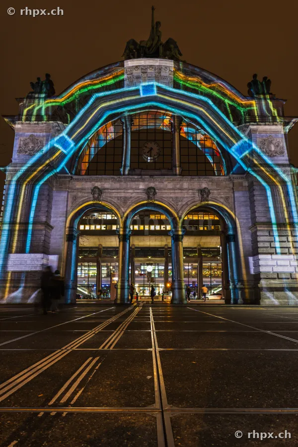 Fassade des Bahnhofs Luzern mit einer farbigen Lichtprojektion des Lilu Lichtfestivals 2024 bei Nacht.