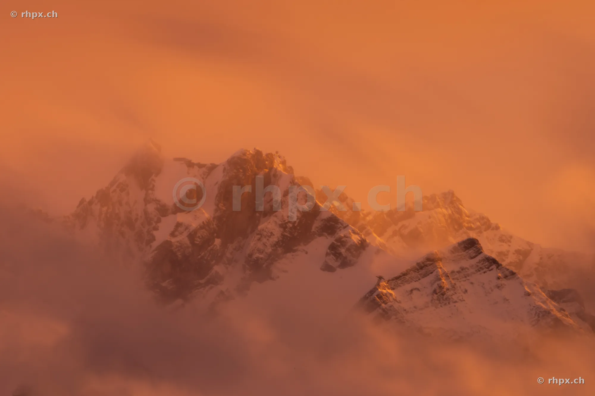 The snow-covered peak of Mount Pilatus in orange evening light surrounded by clouds