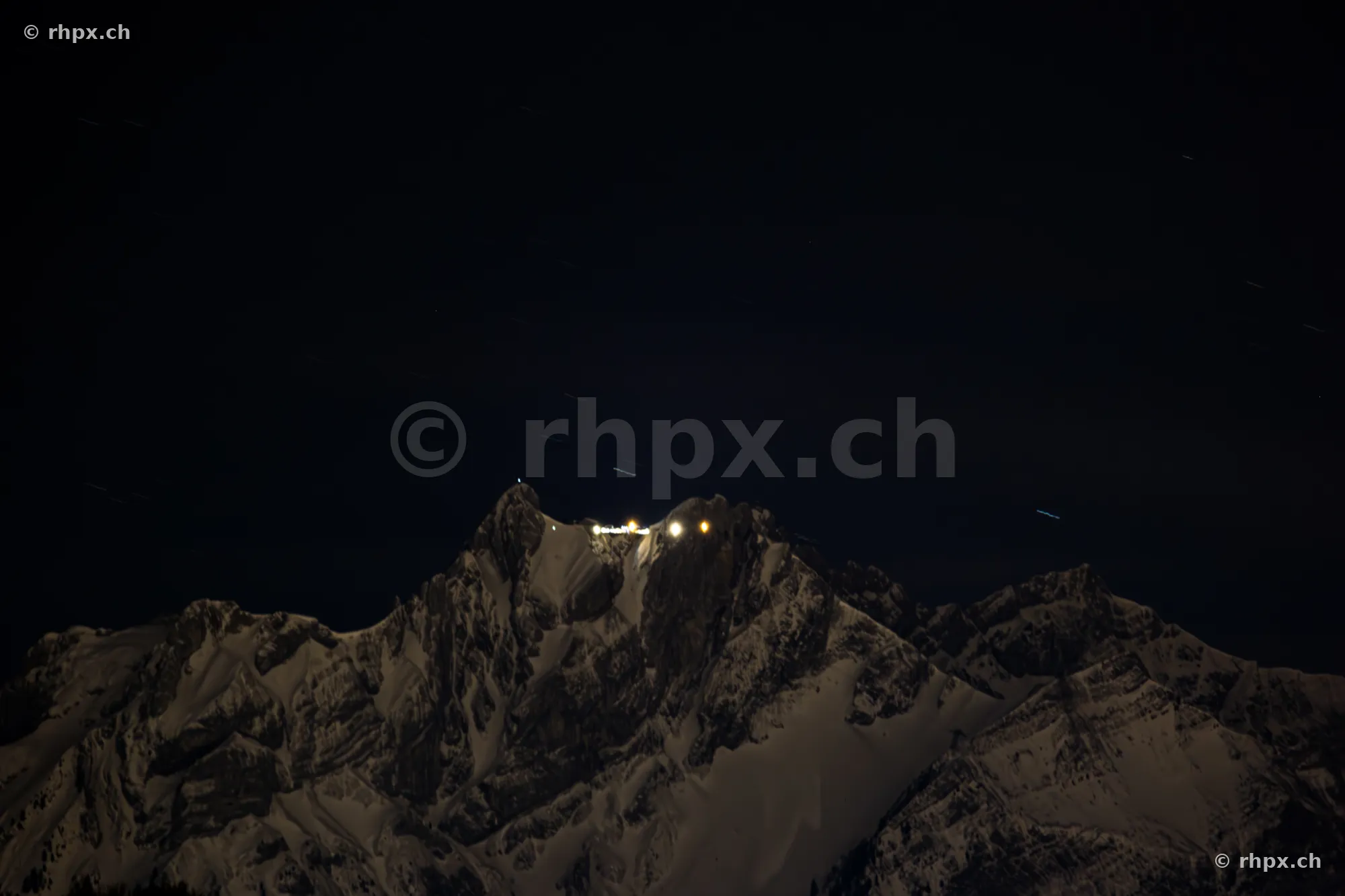 The snow-covered Pilatus summit at night with illuminated buildings under a dark starry sky.