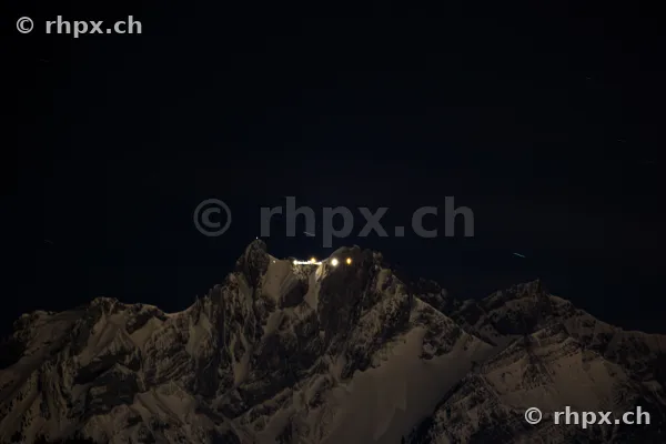 Der schneebedeckte Pilatusgipfel bei Nacht mit beleuchteten Gebäuden unter dunklem Sternenhimmel.