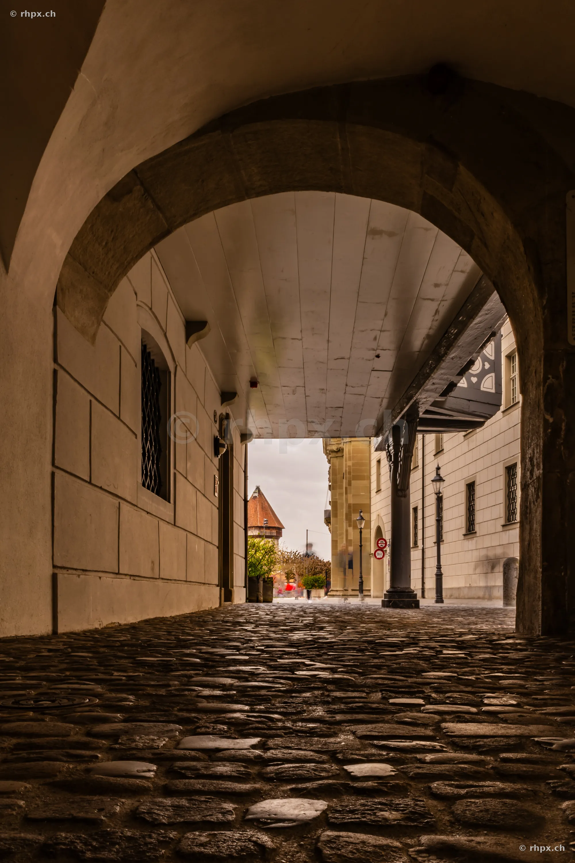 View from a shaded alley with cobblestones towards a historic building and tower in Lucerne.