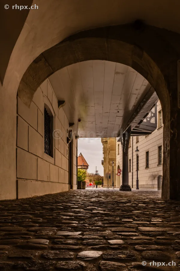 View from a shaded alley with cobblestones towards a historic building and tower in Lucerne.