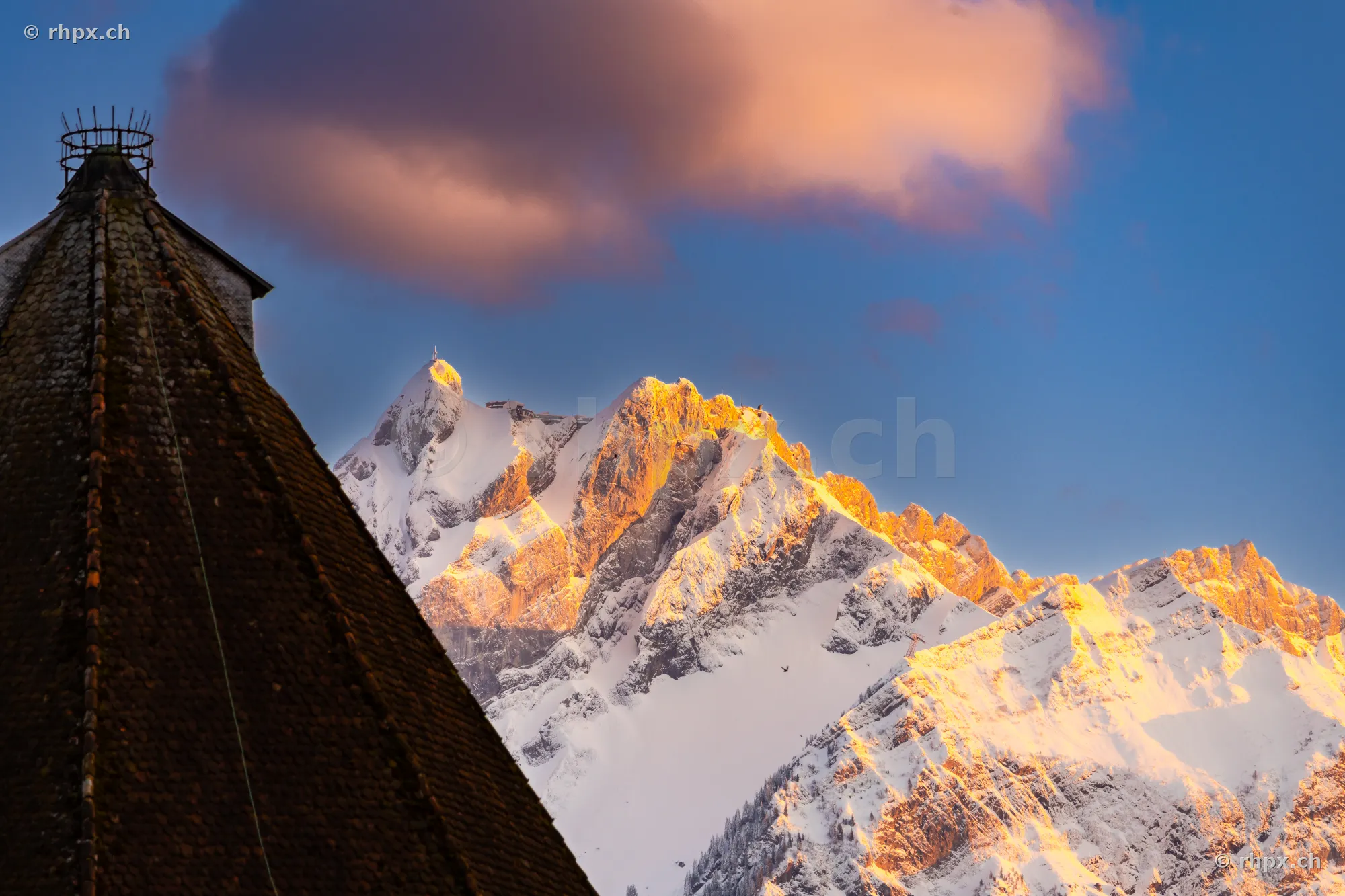 Detailaufnahme einer Kirchturmspitze im Vordergrund mit dem verschneiten Pilatus im Hintergrund bei Abendsonne