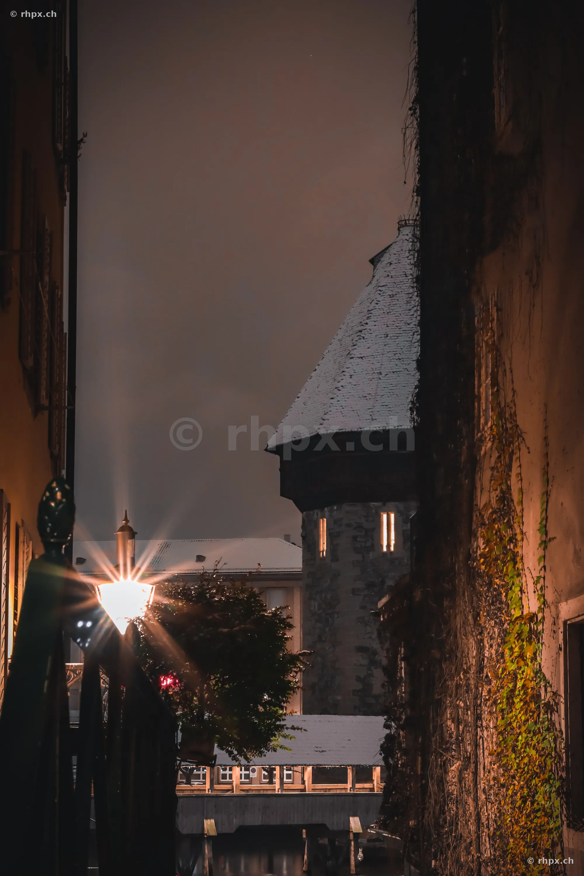 The Wasserturm in Lucerne at night, framed by alley facades and a street lamp.