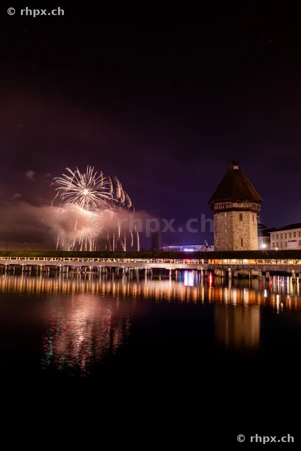 Fireworks in the night sky over the illuminated Chapel Bridge and Water Tower in Lucerne.