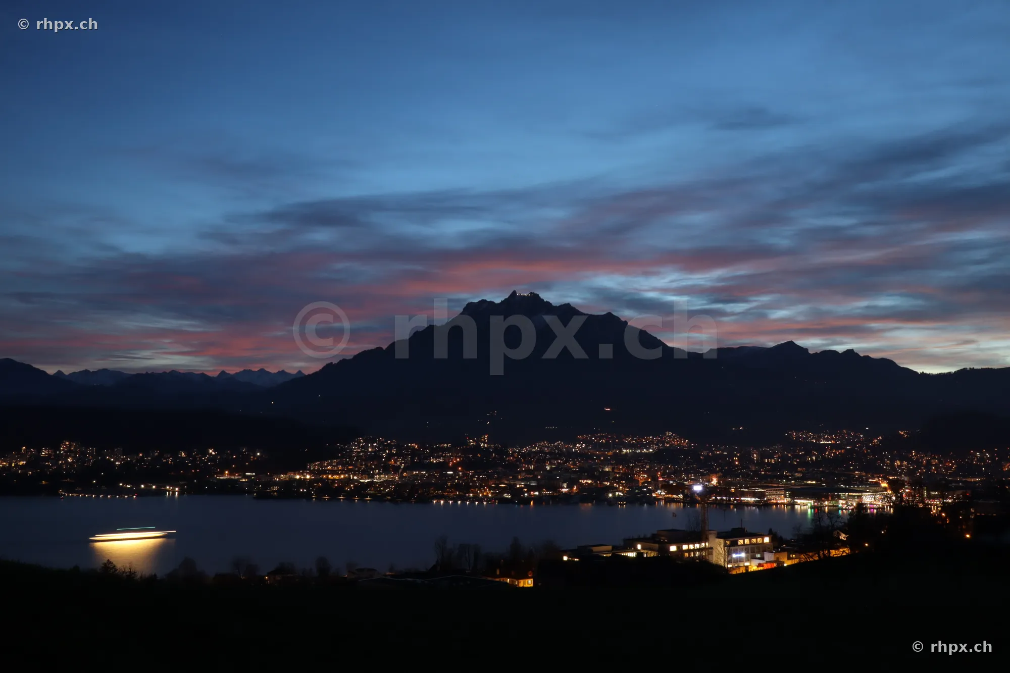 View across Lake Lucerne towards the illuminated city of Lucerne and Mount Pilatus at dusk