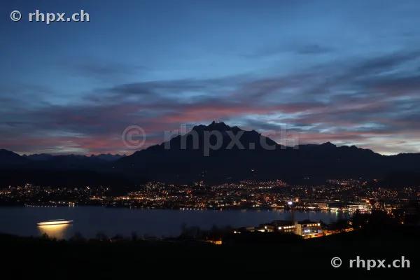 Blick über den Vierwaldstättersee auf das beleuchtete Luzern und den Pilatus in der Abenddämmerung