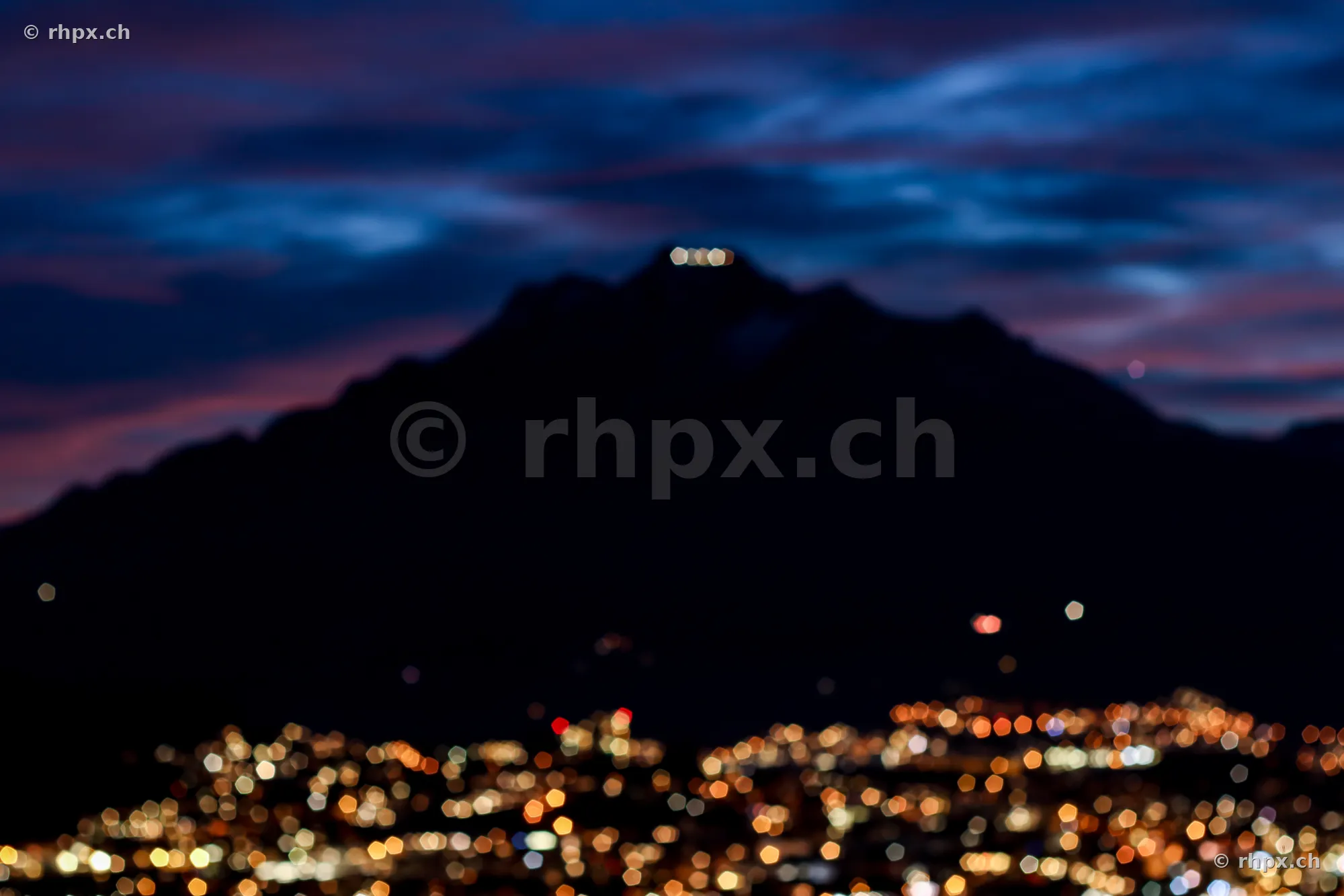 Blurred city lights in front of the dark silhouette of Mount Pilatus at dusk