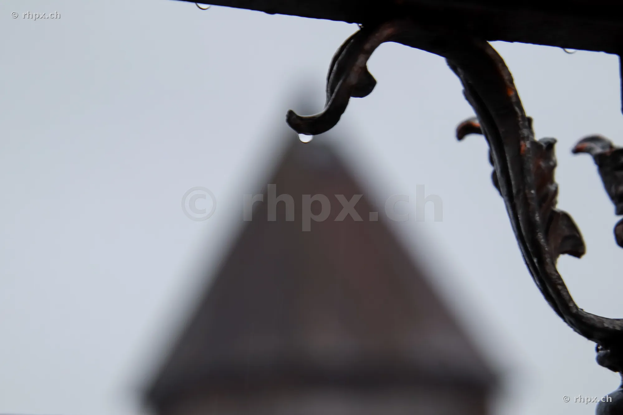 Detailaufnahme von einem Wassertropfen an einem schmiedeeisernen Ornament mit unscharfem Turm im Hintergrund