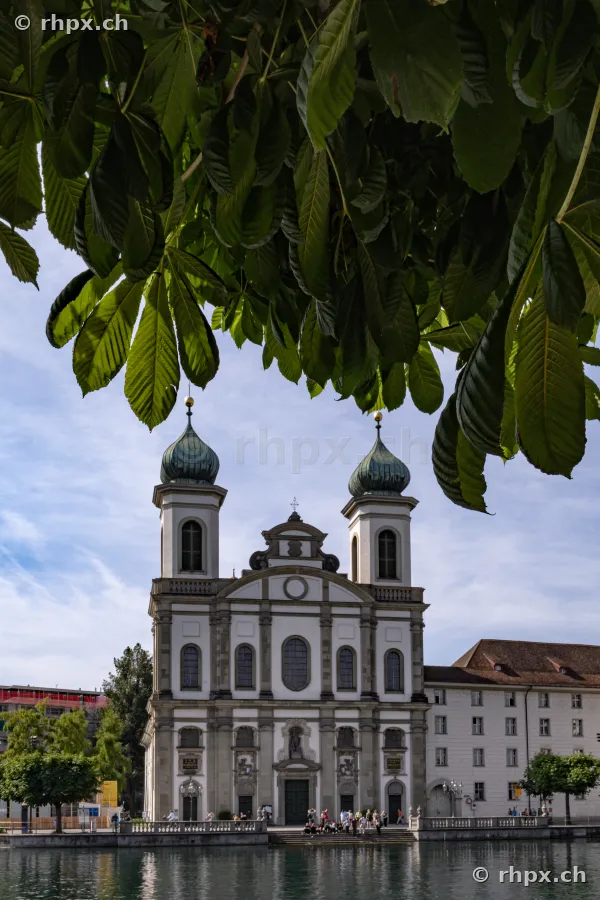 The baroque Jesuit Church in Lucerne seen from the Reuss river, framed by tree branches in the foreground.