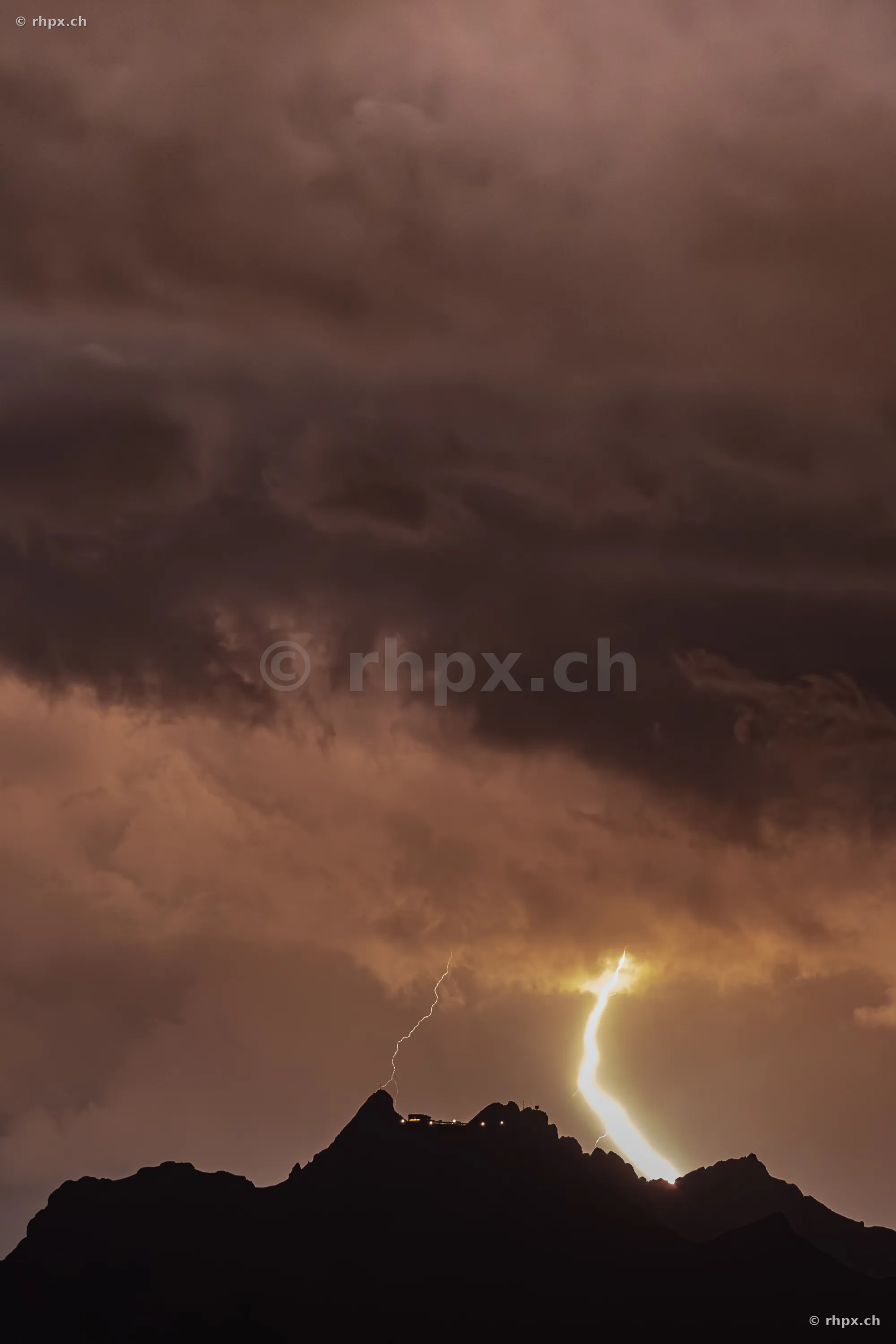 A bright lightning strike discharges over the dark silhouette of the Pilatus summit at night.