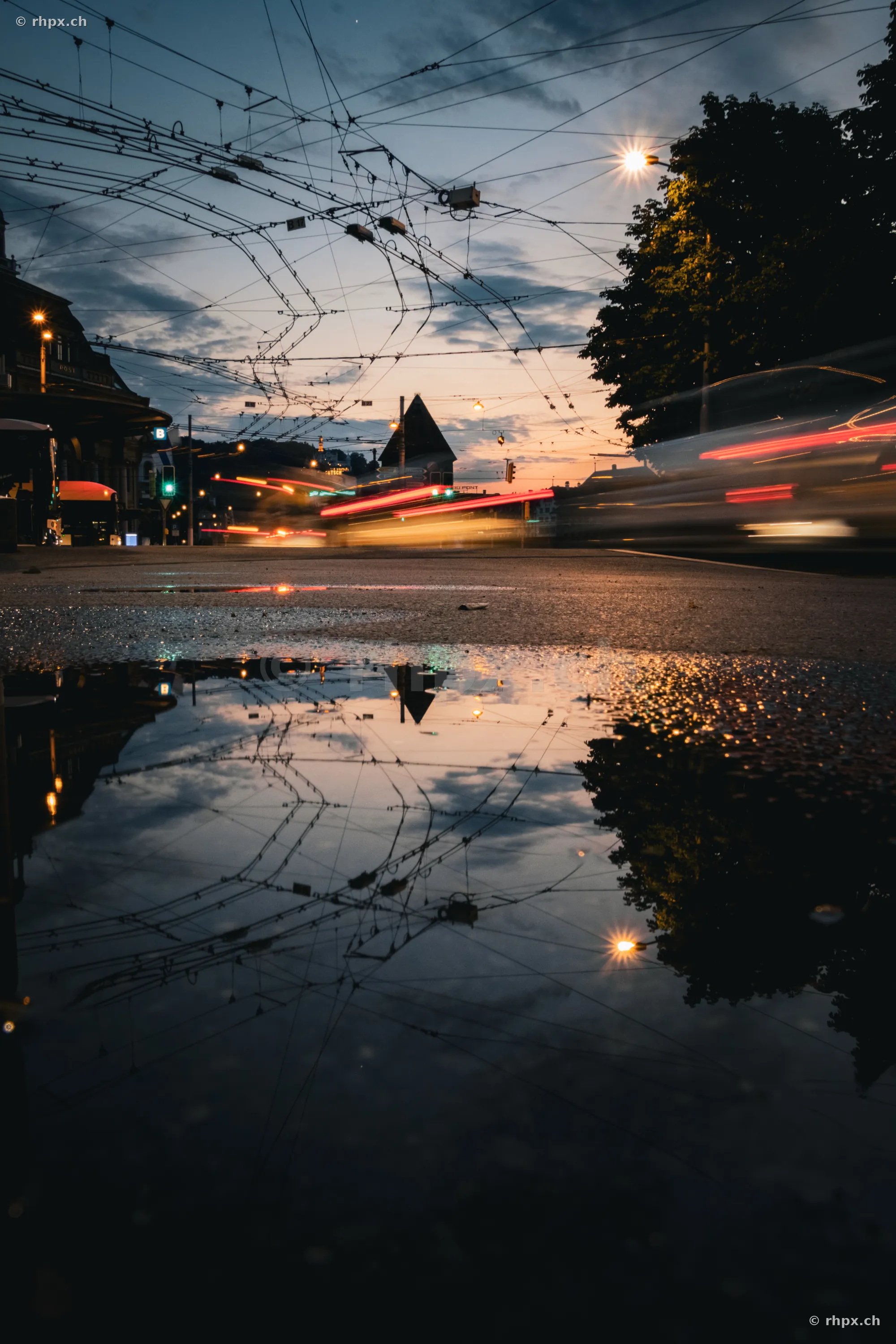 Lichtspuren von Verkehrsmitteln in der Dämmerung auf der Bahnhofstrasse in Luzern mit Spiegelung in einer Pfütze