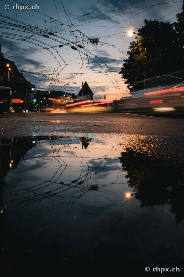 Lichtspuren von Verkehrsmitteln in der Dämmerung auf der Bahnhofstrasse in Luzern mit Spiegelung in einer Pfütze