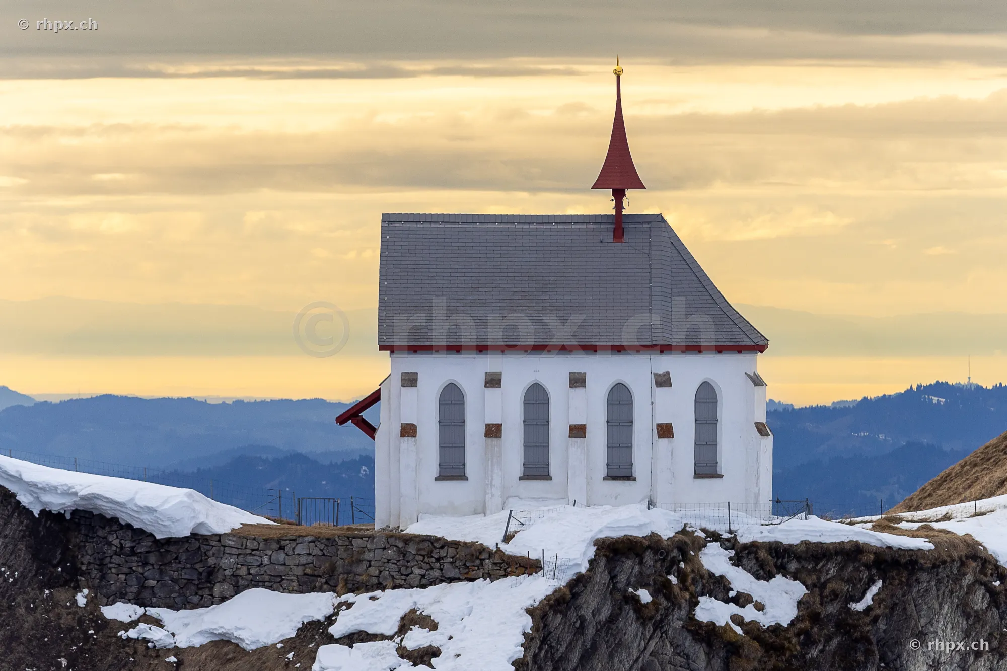 White chapel on the snowy ridge of Mount Pilatus against an evening mountain backdrop.