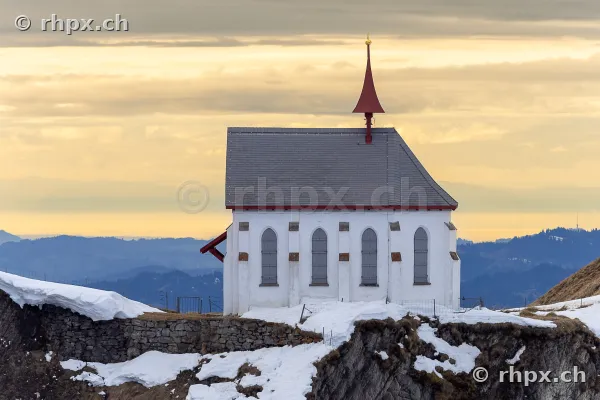 White chapel on the snowy ridge of Mount Pilatus against an evening mountain backdrop.