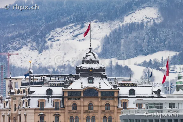 The snow-covered dome and facade of the Hotel Monopol in front of a snow-covered mountain in the background in Lucerne.