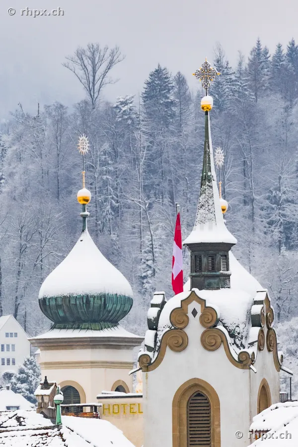 Snow-covered onion domes and part of a hotel building in front of a snowy forest background in Lucerne