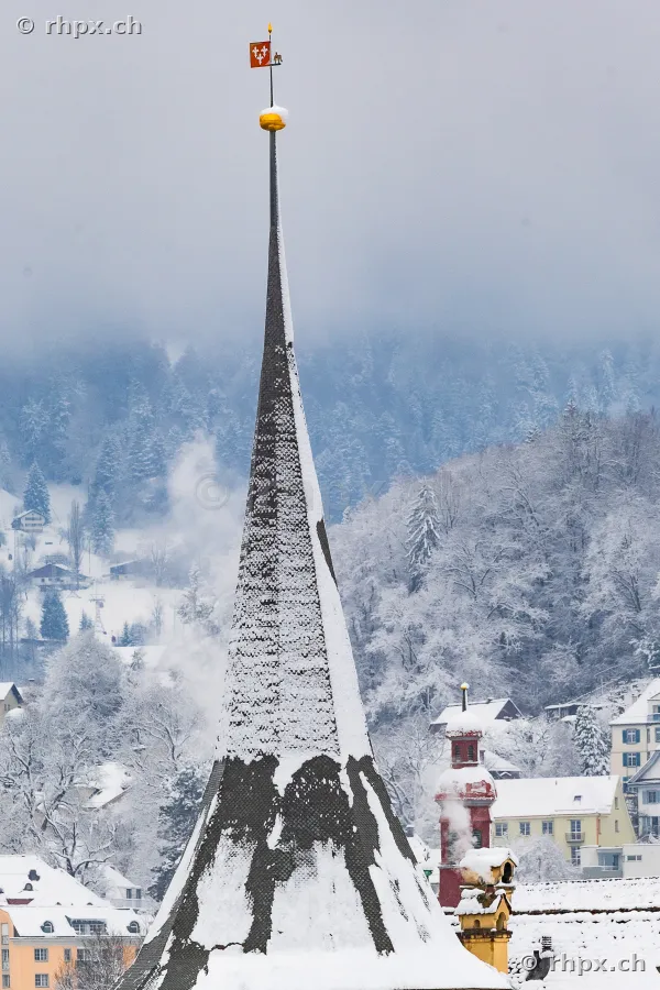 Top of a snow-covered church spire in front of a wooded, snow-covered hill in Lucerne