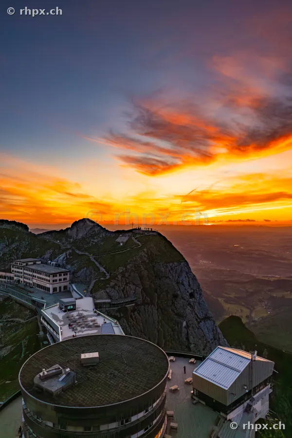 Blick von oben auf die Hotelbauten am Pilatus Kulm während eines orangefarbenen Sonnenuntergangs.