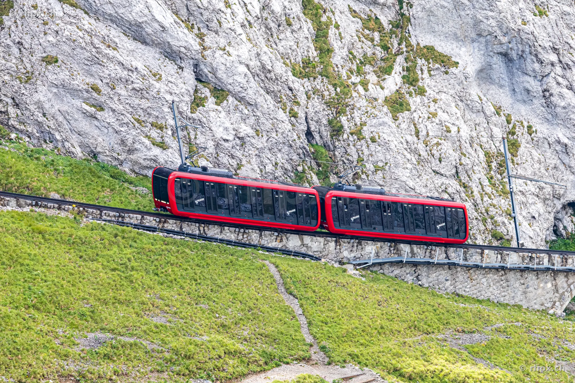 Red cogwheel train driving through steep rock and grass landscape on Mount Pilatus.