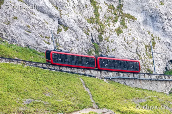 Red cogwheel train driving through steep rock and grass landscape on Mount Pilatus.