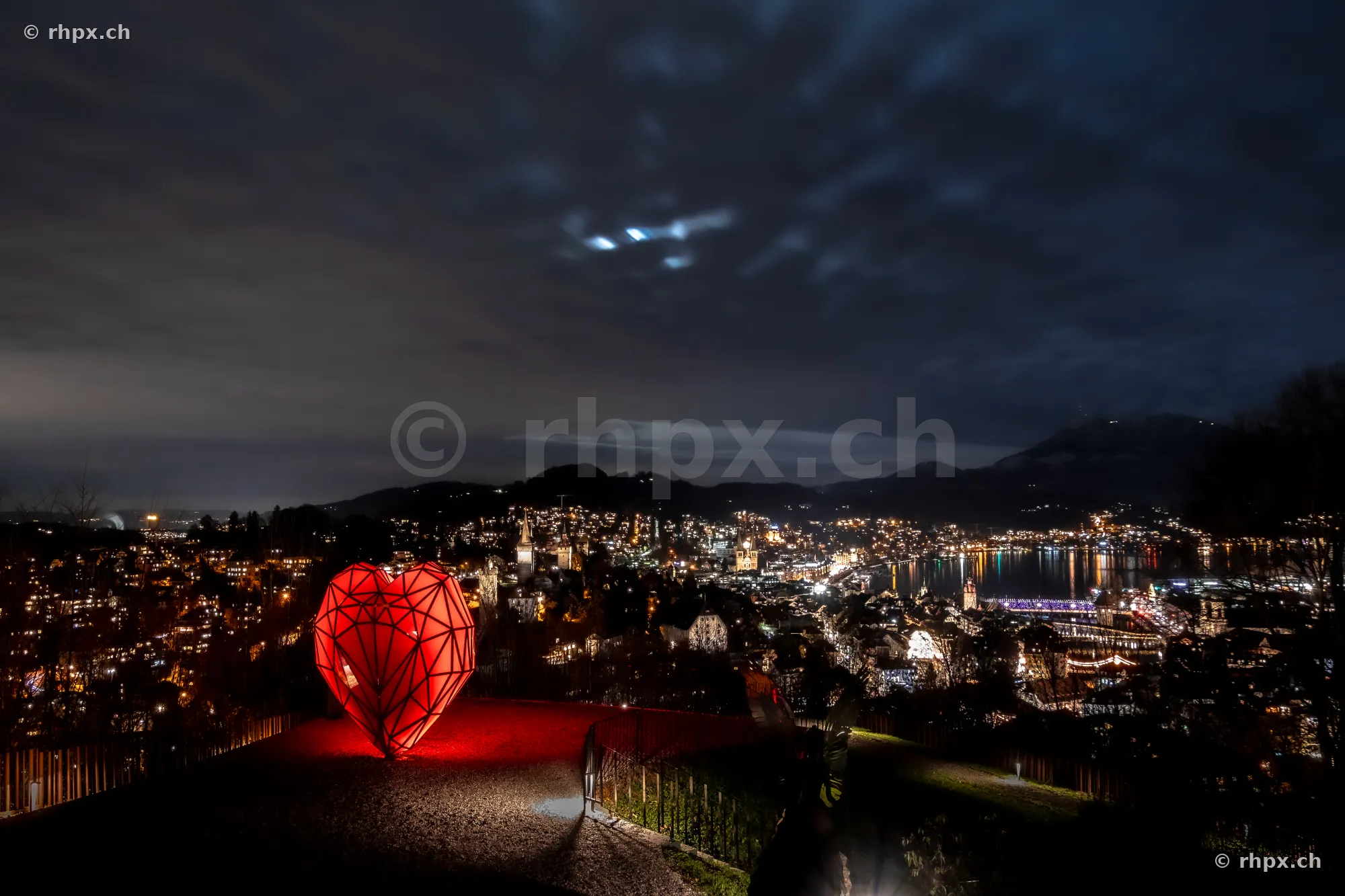A red, illuminated heart object on a viewpoint overlooking the illuminated city of Lucerne at night.