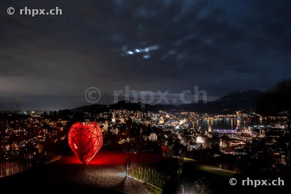 Ein rotes, beleuchtetes Herz-Objekt auf einem Aussichtspunkt oberhalb der nächtlich beleuchteten Stadt Luzern.