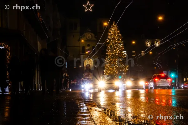 Christmas tree with string lights and star in front of a church in the night-time old town of Lucerne with passing cars.