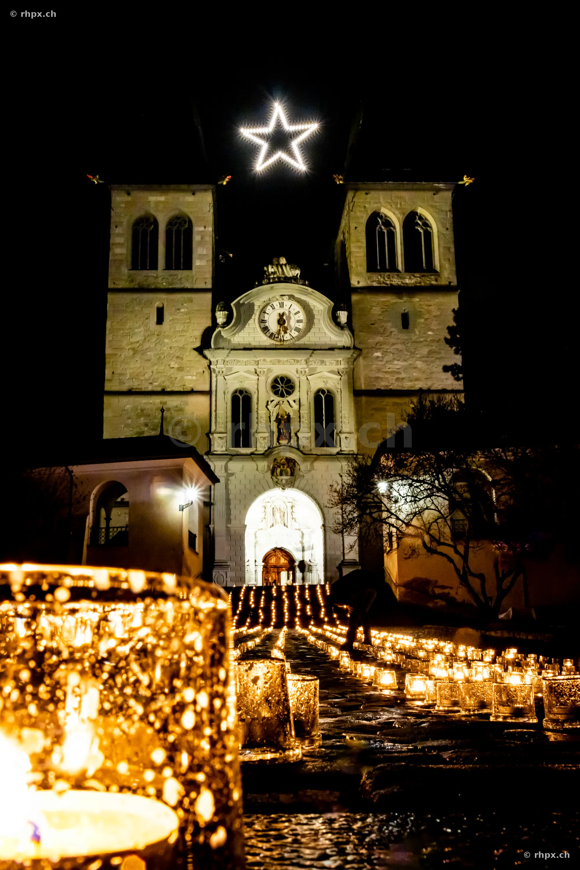 Illuminated Hofkirche in Lucerne with a glowing Christmas star and candles on the forecourt at night.
