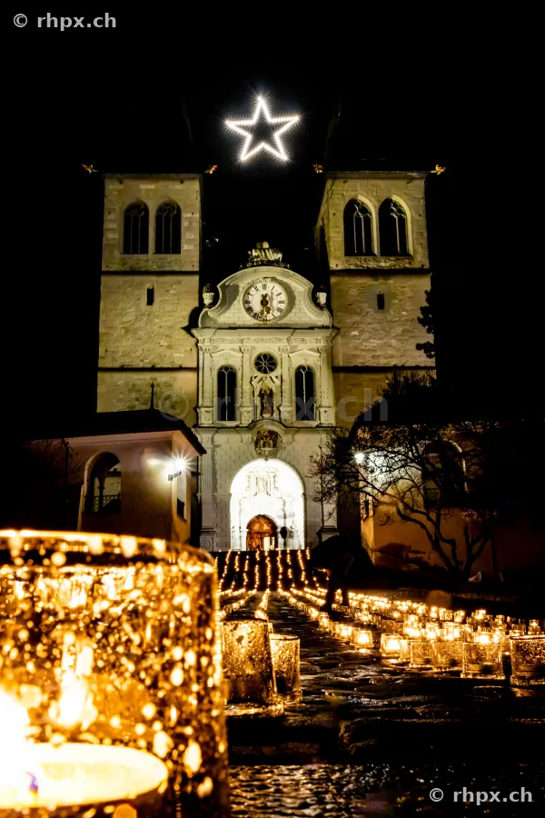 Beleuchtete Hofkirche in Luzern mit einem leuchtenden Weihnachtsstern und Kerzen auf dem Vorplatz bei Nacht.