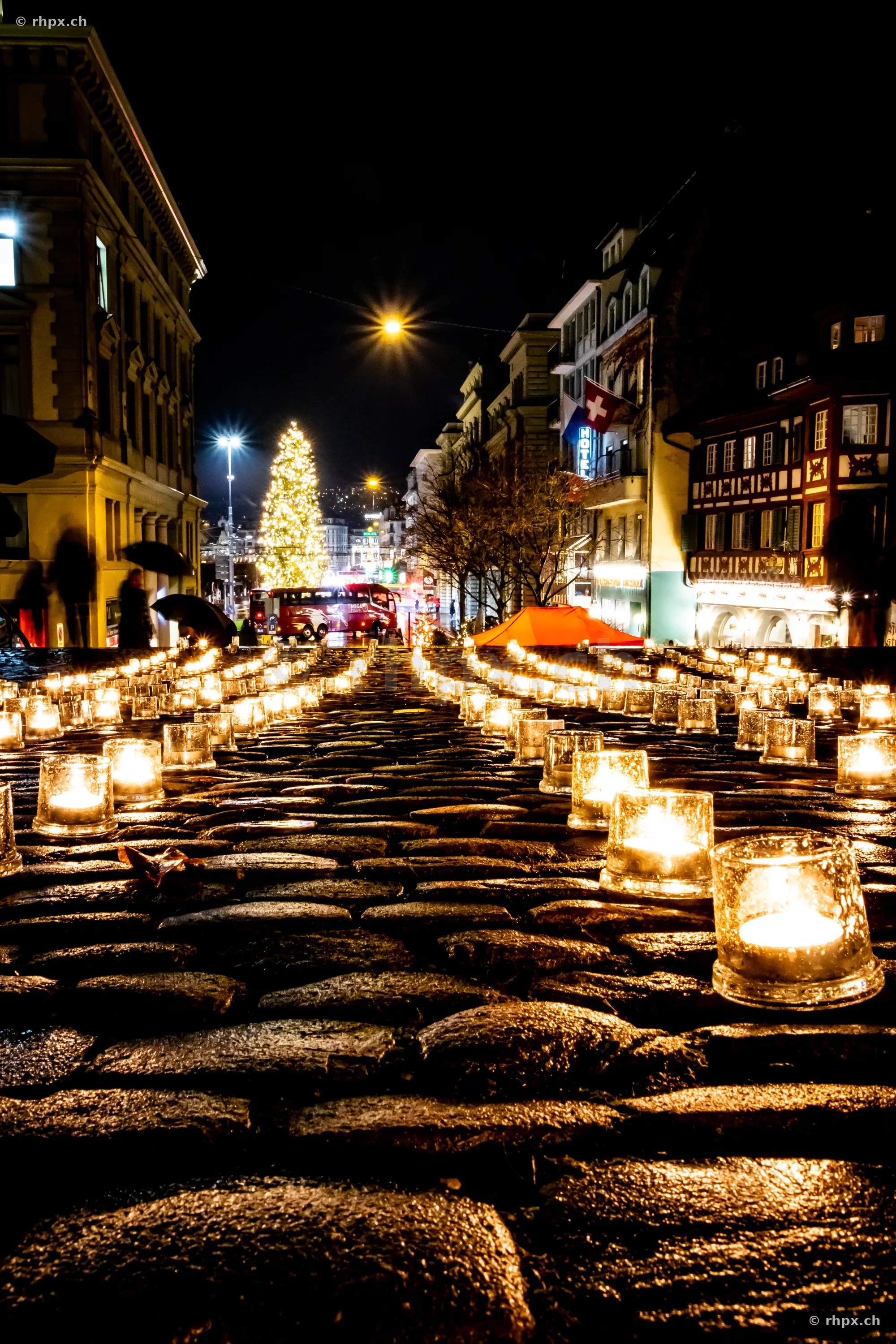 Candles in glass containers on cobblestone pavement in a Christmas-lit street in Lucerne at night.