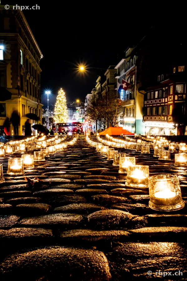 Kerzen in Glasgefässen auf einem Kopfsteinpflaster in einer weihnachtlich beleuchteten Strasse in Luzern bei Nacht.