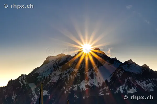 The sun rises directly behind the jagged peak of the snow-covered Pilatus mountain.
