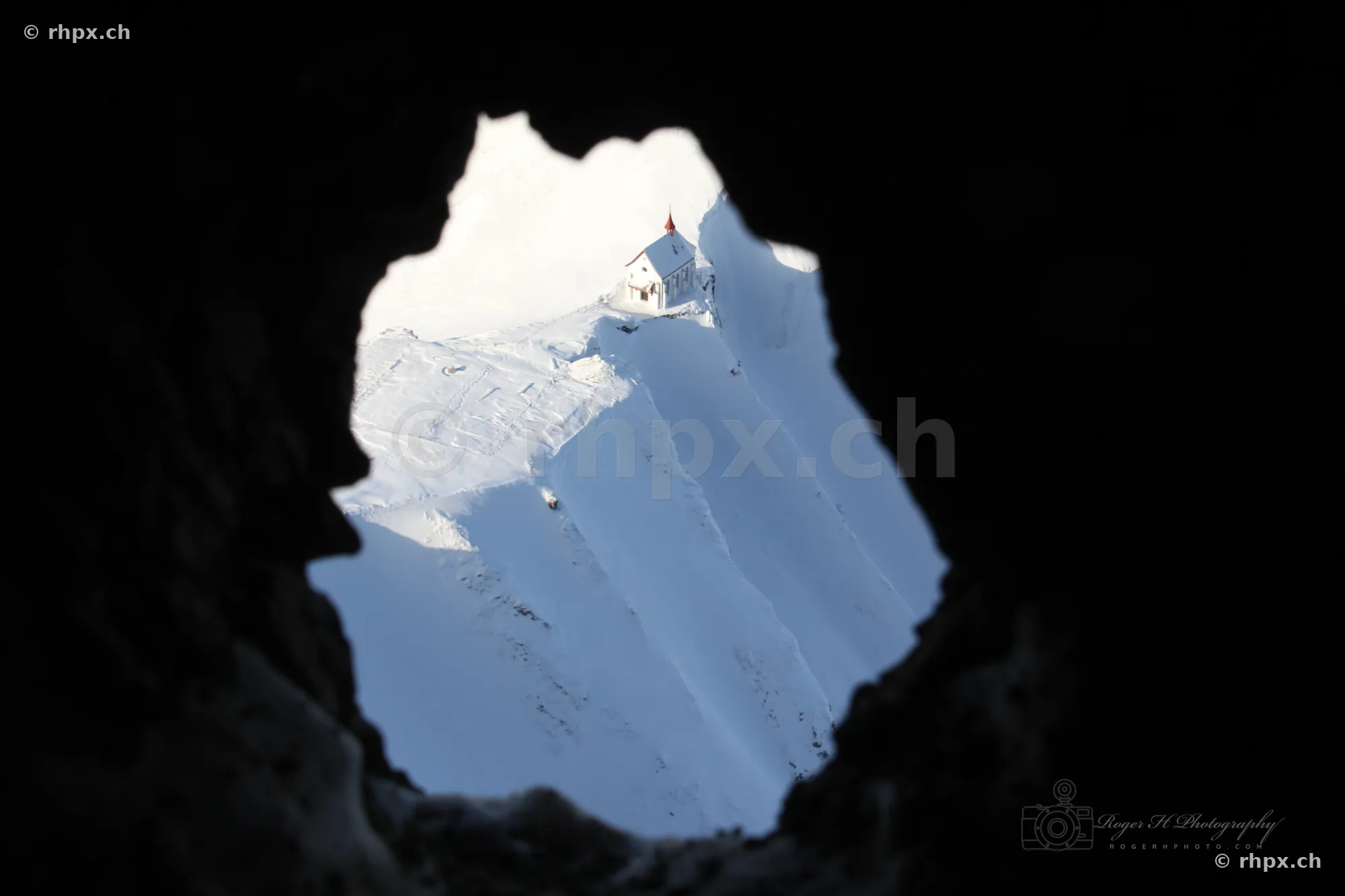 View from a dark rock opening towards the snow-covered Pilatus chapel in bright daylight.