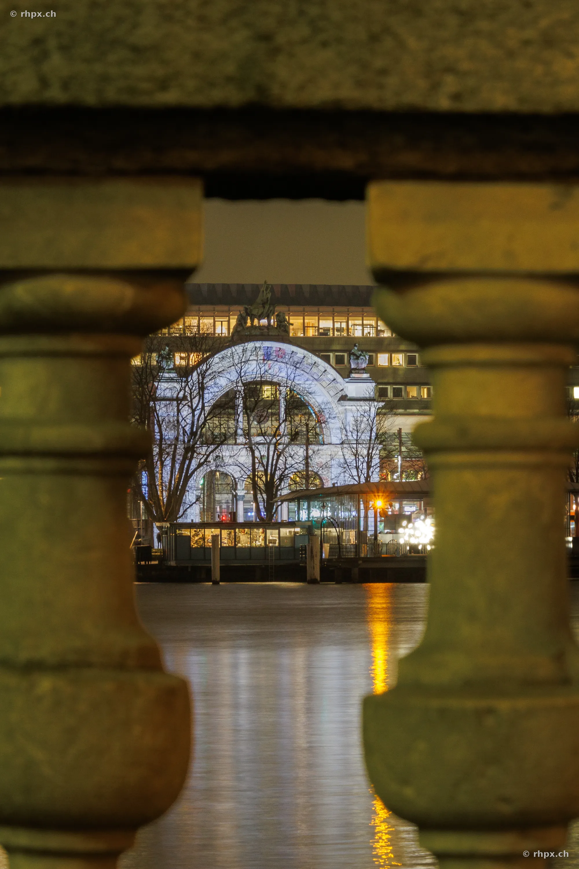 View through a stone balustrade of the illuminated Lucerne train station during the 2024 Lilu Light Festival.