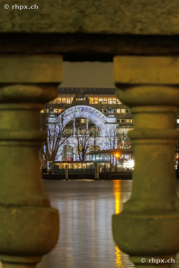 Blick durch eine steinerne Balustrade auf den beleuchteten Bahnhof Luzern während des Lilu Lichtfestivals 2024.